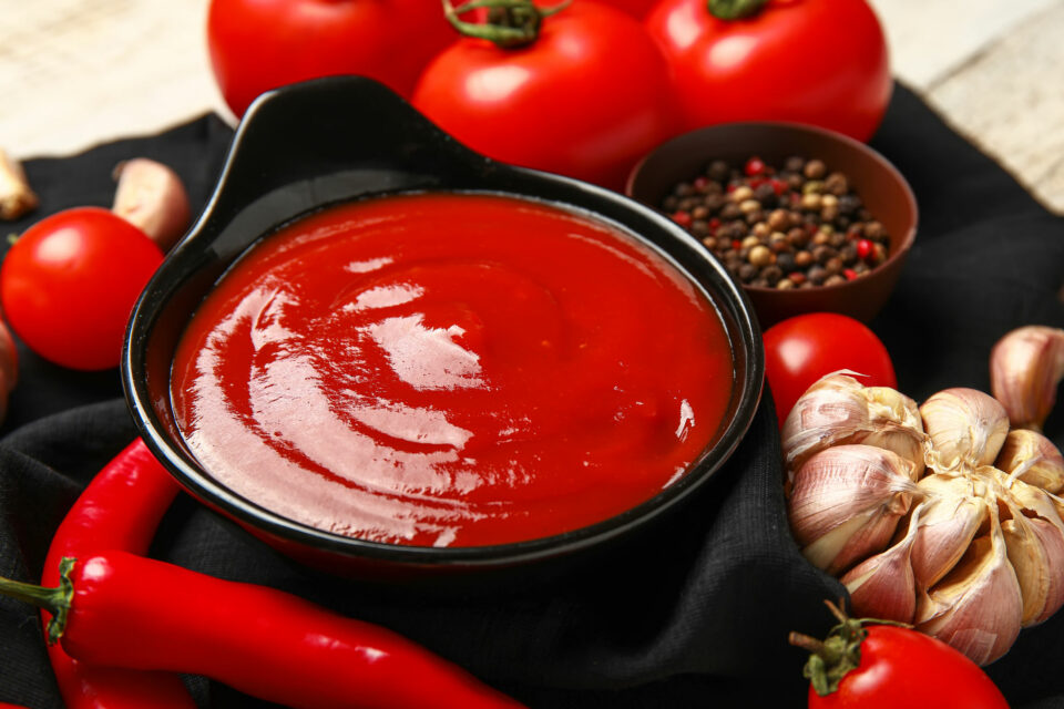 Ketchup or tomato sauce in a black ceramic bowl. Tomatoes, chili and garlic can be seen in the background